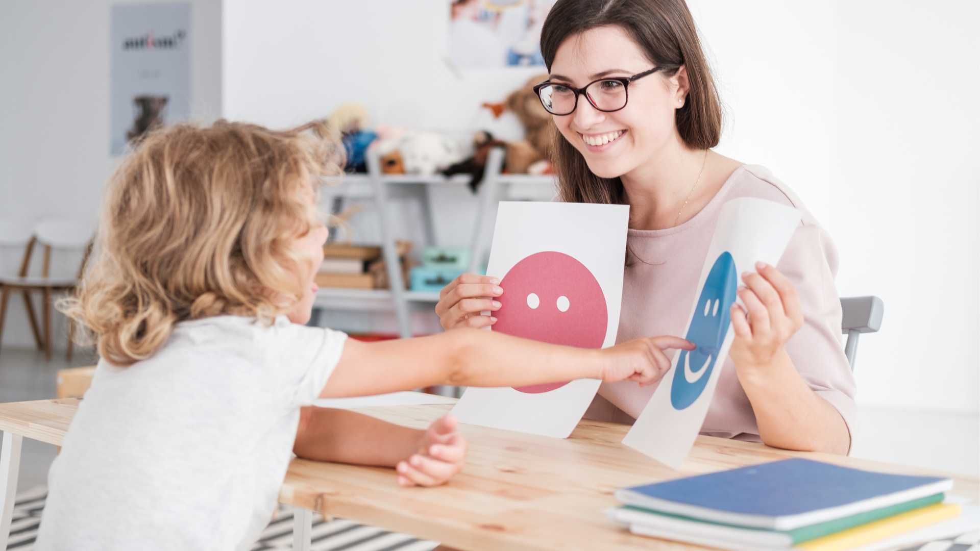 Autism support groups in Pennsylvania: At a table, a woman is holding up two papers, one with a red face and one with a smiling blue face. A child points to the smiling one.