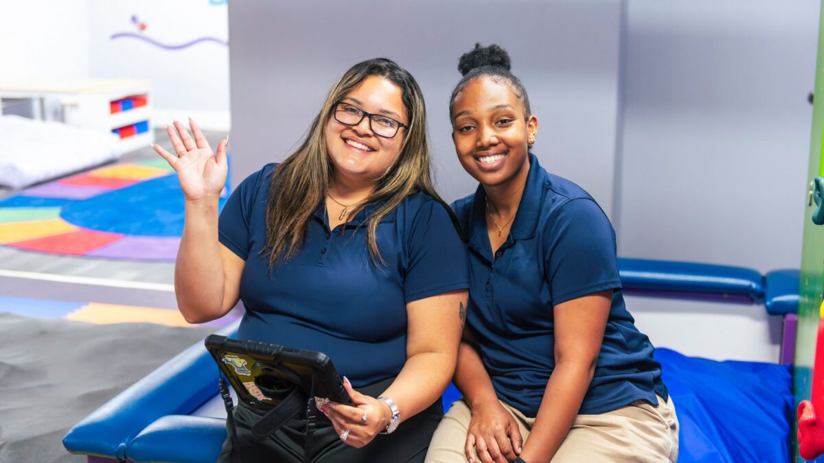 Two women sit together on a couch in a playroom as an autism support team