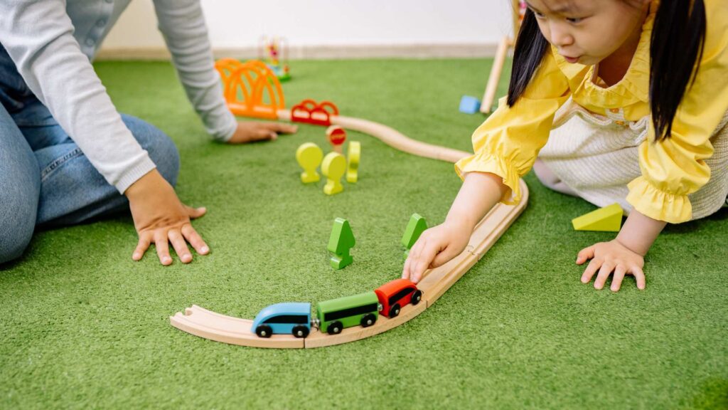 A child plays with a wooden train set on a green mat next to an adult, showing a calm moment that relates to an autism diagnosis and how families can support children with ASD at home.